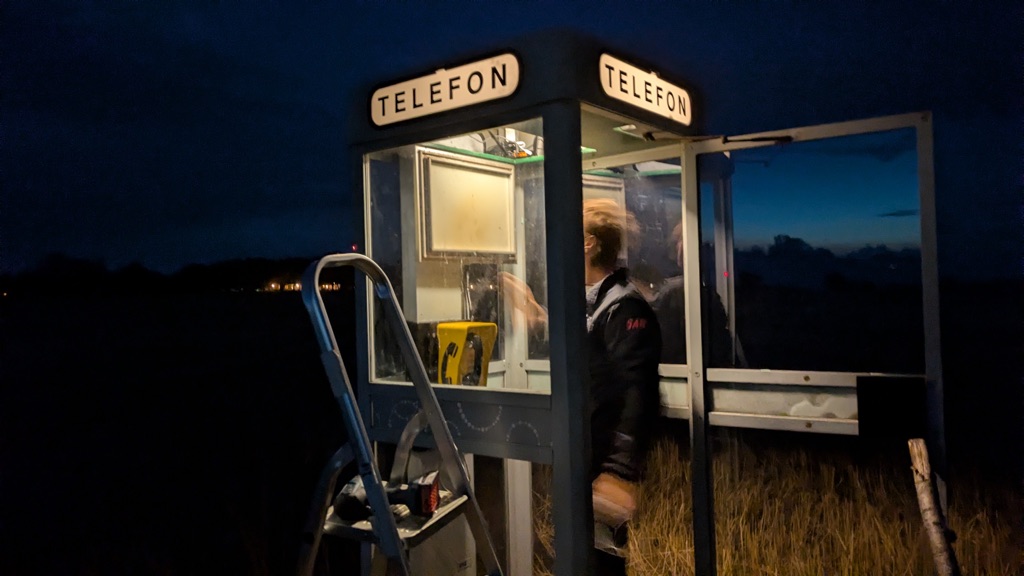 Two people at the lit booth at dusk, with a ladder and a drill